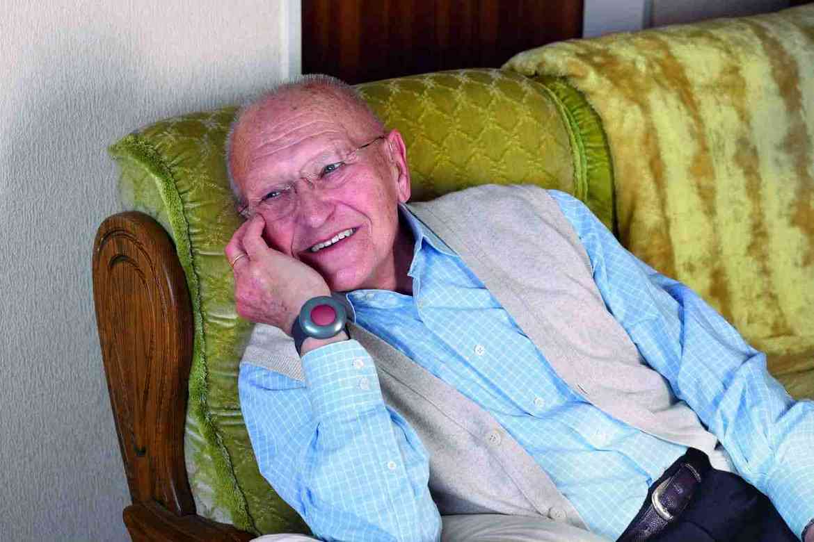 Ein älterer Herr sitzt auf seinem Sofa. Ums Handgelenk trägt er den Hausnotrufknopf des ASB.